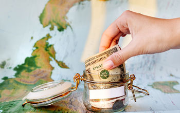 Collecting money for travel. Glass tin as moneybox with cash (Photo via Aksenovko / iStock / Getty Images Plus)